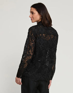 A woman with dark hair stands against a plain background, wearing the HINSON WU - MARGOT LACE SHIRT JACKET, featuring intricate lace detailing and satin trim. She has elegantly paired it with dark pants, her profile turned slightly sideways as she offers a subtle smile.