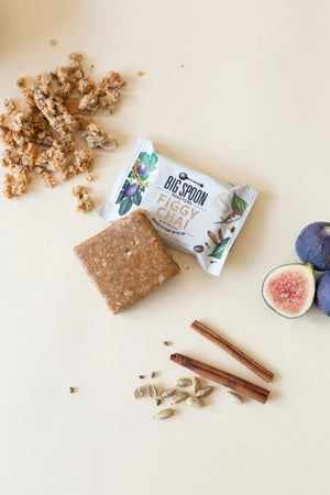 The image displays the packaging of a "BIG SPOON ROASTERS - FIGGY CHAI NUT BUTTER SNACK BAR." The white wrapper is adorned with illustrations of figs, spices, and nuts, emphasizing that it contains gluten-free whole grains and non-GMO pea protein. Additionally, it is free from dairy, gluten, soy, and palm oil, with a weight of 60g (2.1oz).