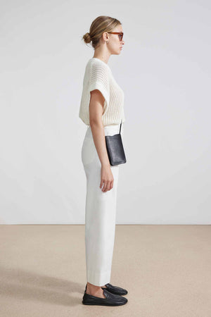 A person with hair in a bun is facing away, wearing sunglasses, a cream knit top, and the APIECE APART - CLASSIC MERIDA PANT that creates a sleek silhouette. Black shoes and a thin black crossbody strap complete the look against a light, minimalist setting.