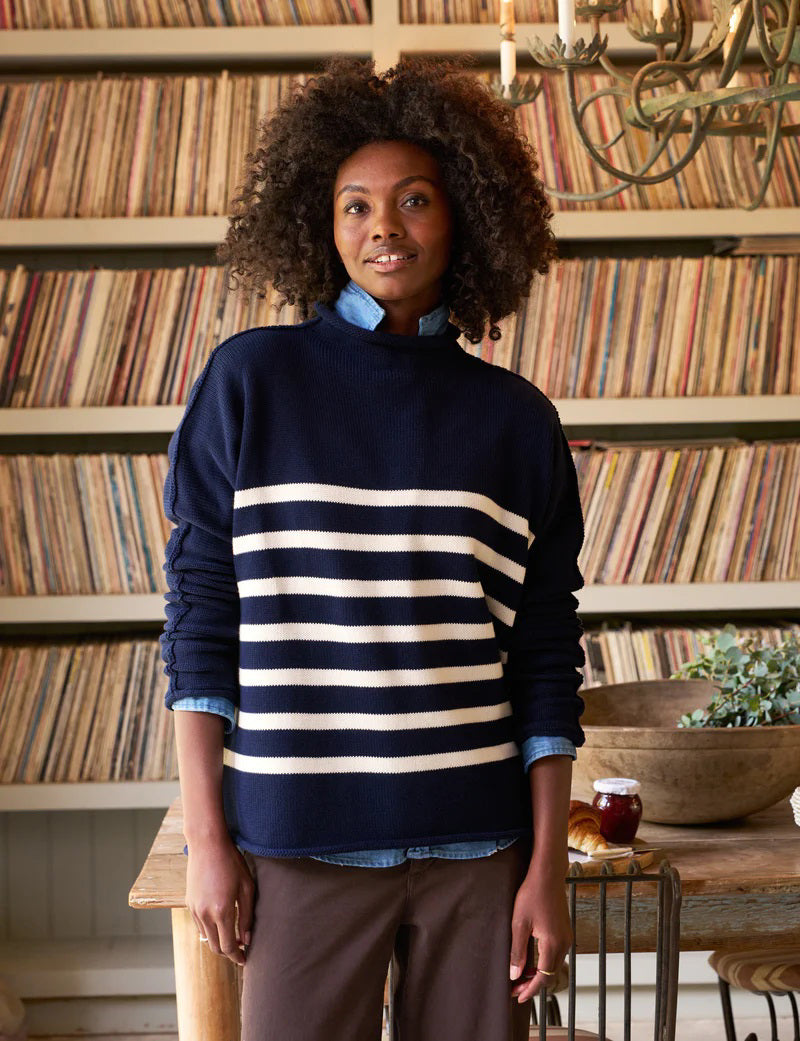 A person with curly hair stands indoors, wearing a Frank & Eileen Monterey Rolled Funnel Neck Sweater in Navy Cream Stripe with a blue shirt underneath. They are in front of a wooden table adorned with plants, a candle, and a bowl, while shelves filled with vinyl records are visible in the background.