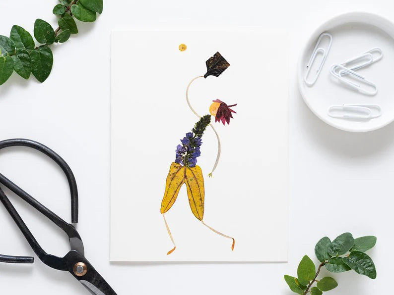 The PICKLEBALL GREETING CARD by PETAL PEOPLE PRESS features a whimsical piece of botanical art depicting a figure joyfully tossing an object resembling a hat. Crafted from pressed flowers, leaves, and plant parts, the design is set against a white backdrop with scattered office supplies and greenery adding to its enchanting appeal.