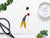 The PICKLEBALL GREETING CARD by PETAL PEOPLE PRESS features a whimsical piece of botanical art depicting a figure joyfully tossing an object resembling a hat. Crafted from pressed flowers, leaves, and plant parts, the design is set against a white backdrop with scattered office supplies and greenery adding to its enchanting appeal.