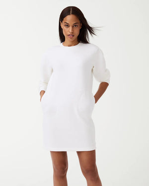 A woman is wearing the SPANX AirEssentials Crew Neck Dress, a black, long-sleeve, knee-length dress from the SPANX brand, with her hands in the functional pockets. She is looking to the side and has long, straight hair. Paired with white sneakers, she stands against a plain white background in this light-as-air fabric attire.