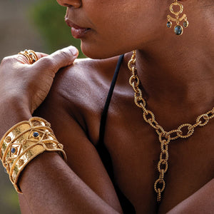 Close-up of a person wearing intricate 18K gold plated jewelry, including a Small Rope Link Necklace, dangly earrings, and bracelets with gemstone accents. The person is resting their hand on their shoulder, showcasing the detailed design of the accessories and a CAPUCINE DE WULF - VICTORIA SMALL CHAIN NECKLACE.