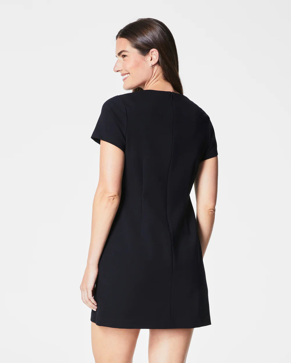 A person with long hair is wearing the figure-flattering SPANX - PONTE MINI DRESS in black, paired with silver heels, standing against a plain white background. They gaze ahead with a neutral expression, one hand slightly raised.