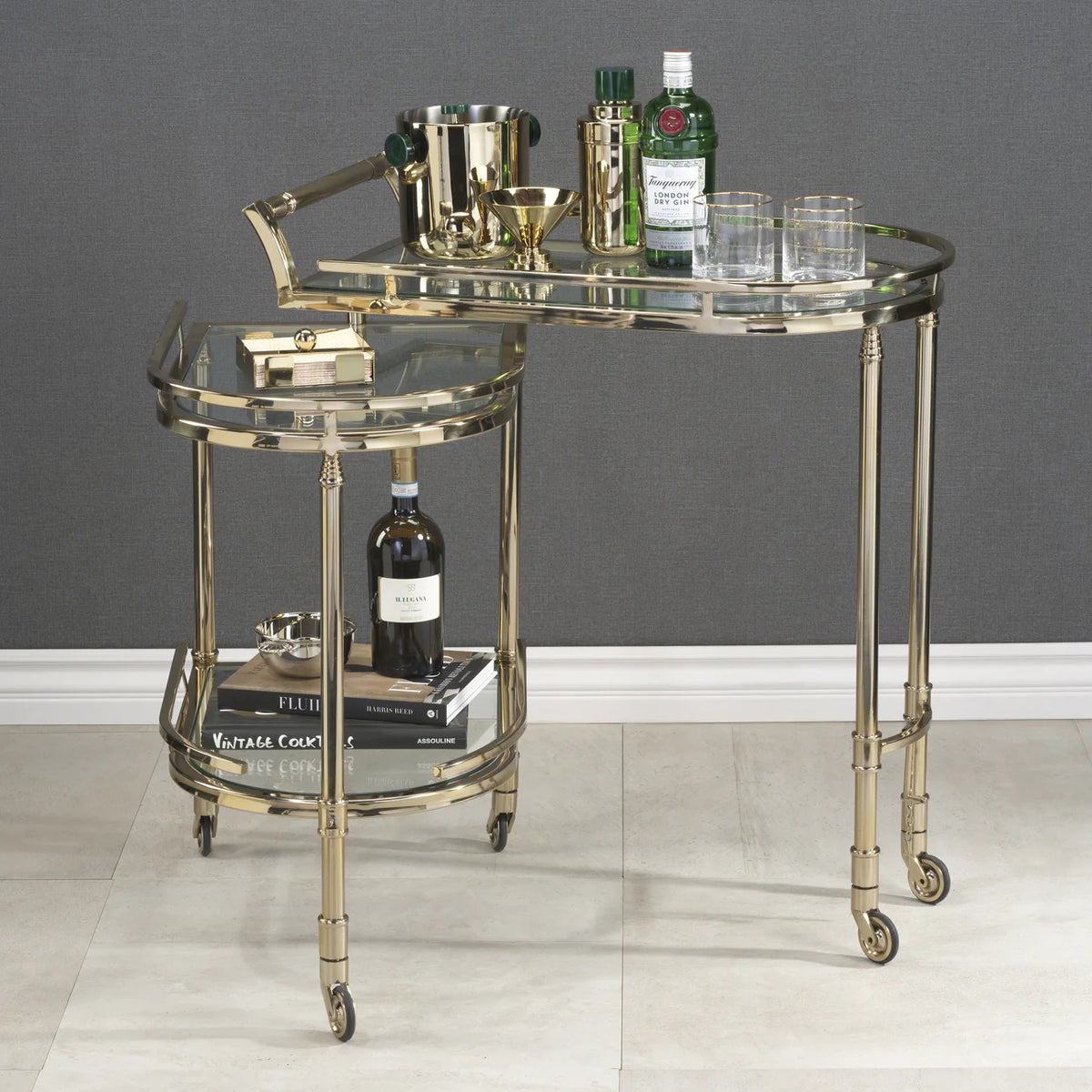 The SPIGA GOLD BAR CART by ZODAX features a gold alloy glass frame, two glass shelves, and four small wheels for easy mobility and stylish serving.