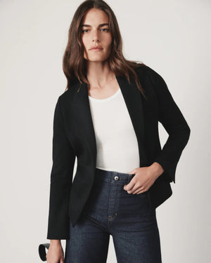 A person with long hair poses confidently, wearing the SPANX - PONTE BLAZER in a versatile black hue, crafted from premium ponte fabric, layered over a white top and dark jeans. The plain, light-colored background creates a minimalist setting.