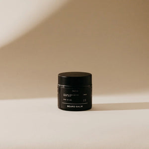 A small black container of PRESTON's cruelty-free beard balm is placed against a neutral, softly-lit backdrop. The label reads "PRESTON - BEARD BALM" along with other details in light/white text. Shadows fall to the right, adding depth to the minimalist design setting that showcases its satin finish.