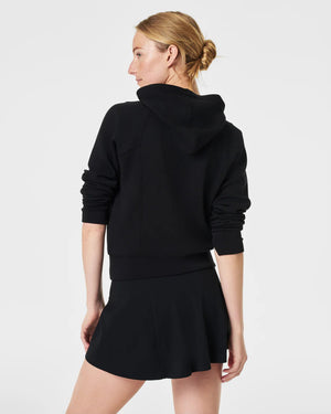 A person with blonde hair is wearing the SPANX AirEssentials Full Zip Hoodie made from ultra-soft fabric, with their hands in the pockets and a black skirt. They are standing against a plain, light-colored background and smiling softly.