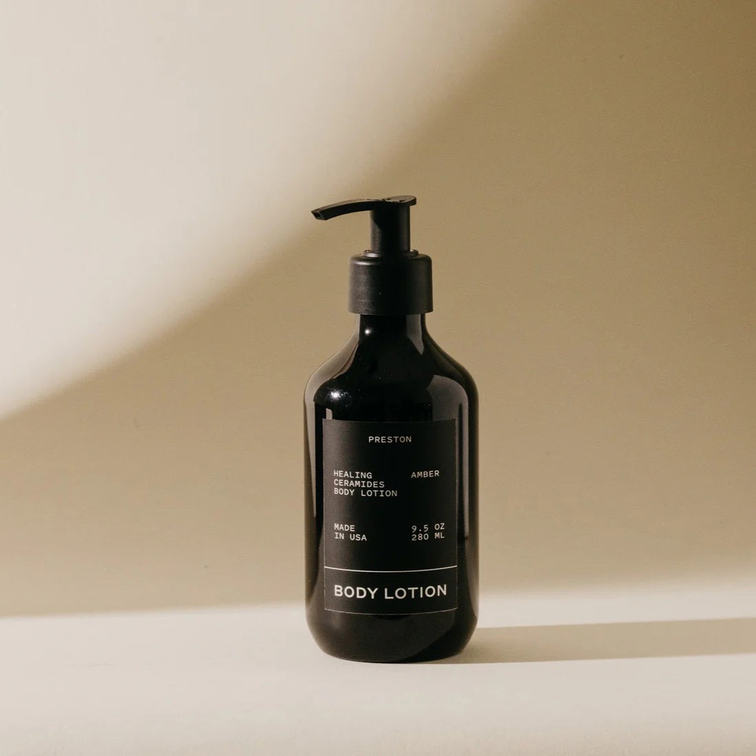 A sleek, dark brown bottle of Preston Body Lotion with a pump dispenser is displayed against a neutral beige background. The label reads &quot;PRESTON,&quot; &quot;Healing Amber Body Lotion,&quot; &quot;Made in USA,&quot; and &quot;9.5 fl oz / 266 ml.&quot; Infused with plant-based moisturizers, its natural formula is accentuated by soft lighting that casts shadows to the right of the bottle.
