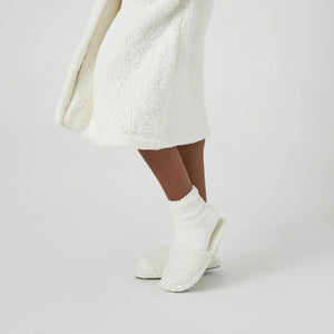 A person stands on a white background, wearing a cozy, textured white robe, KASHWERE - ADULT SOCKS by KASHWERE, and white slippers. Only the lower half of the body is visible. The attire appears ultra-plush and comfortable, ideal for lounging or relaxation.