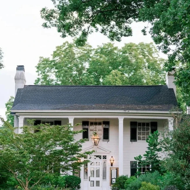A white two-story house with black shutters, a dark roof, and a central lantern above the front door, surrounded by lush green trees and shrubs.