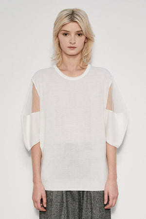 A person with shoulder-length blonde hair stands against a plain background, exuding an air of whimsy. They are wearing the MEIMEIJ - KNIT TOP WITH SHEER SLEEVES by MEIMEIJ and loose gray pants. Their expression is neutral.