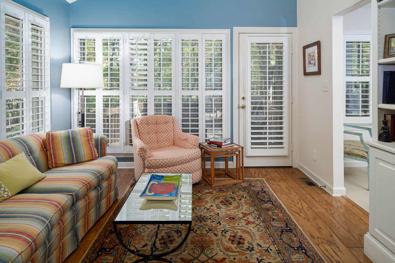 A cozy living room with a striped sofa, patterned armchair, glass coffee table, and a colorful rug. Large windows with white shutters let in natural light. Walls are blue and white, and hardwood floors add warmth.