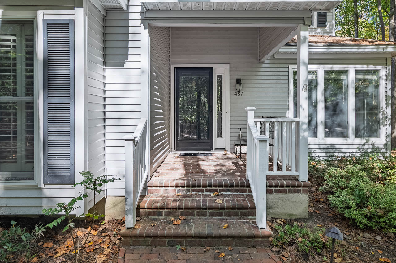 A small brick porch with white railings and steps leads to a glass front door on a light gray house, surrounded by green shrubs and trees. Sunlight filters through the trees, casting shadows.