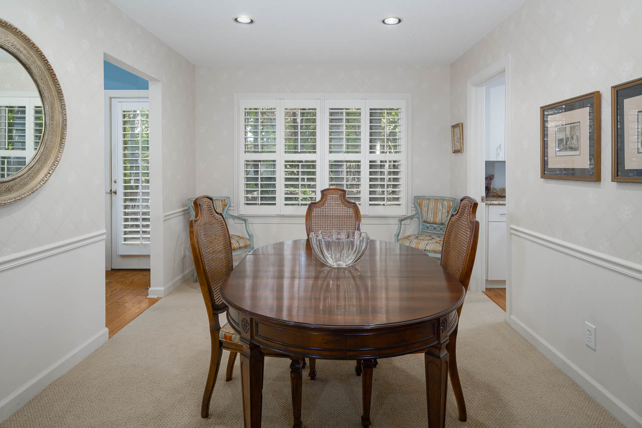 A dining room with a round wooden table, four chairs, a glass bowl centerpiece, beige carpet, white walls, two armchairs near shuttered windows, a large mirror, and framed pictures on the wall.