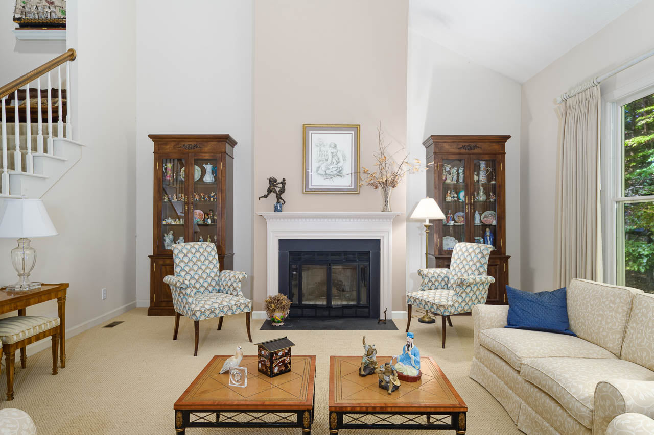 A bright, elegant living room with a white fireplace centered between two glass cabinets. Patterned chairs, a cream sofa, and wooden coffee tables sit atop a beige carpet. Natural light streams through large windows.
