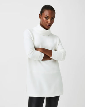 Dressed in the luxurious comfort of a SPANX AireEssentials Turtleneck Tunic, characterized by its black hue and lightweight fabric, a person stands with their back to the camera. Their elegantly tied up hair complements the matching black pants against a plain white backdrop.