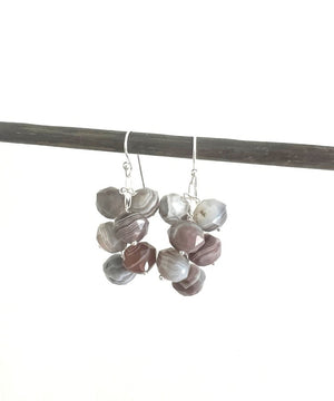 A pair of ANN LIGHTFOOT - Faceted 9mm Botswana Agate Droplet Earrings features clusters of gray and brown beads, creating a textured look. They hang from a dark rod against a plain white background.