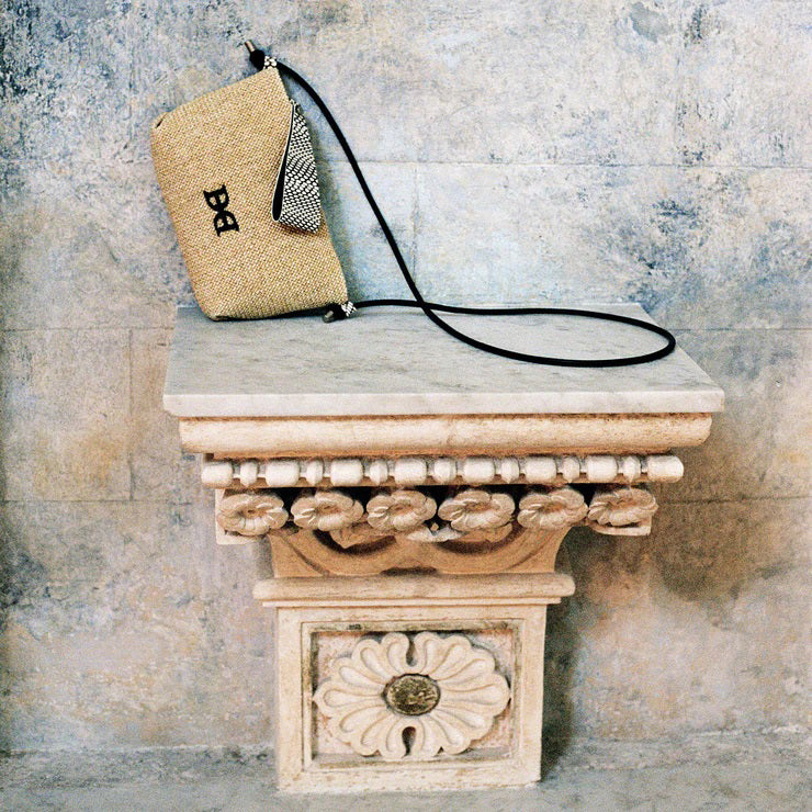 The DANIELLA LEHAVI - VEGA STRAW CLUTCH, featuring a beige woven design and an adjustable black strap, elegantly rests on an ornate stone pedestal adorned with intricate carved designs. The backdrop showcases a textured stone wall.