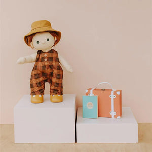 A Dinkum Doll from Olli Ella USA's Travel Togs collection, featuring dark hair and dressed in a denim jacket, mustard pants, and black shoes, stands on a wooden surface. Beside the doll is a teal See-Ya Suitcase with white trim and hardware. In the background, another larger teal suitcase is partially visible.