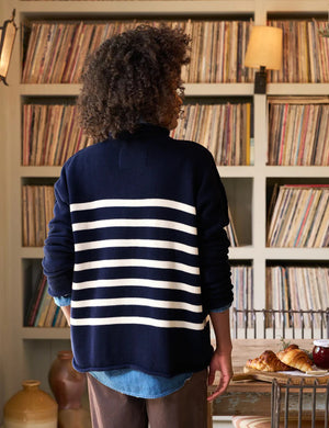 A person with curly hair stands indoors, wearing a Frank & Eileen Monterey Rolled Funnel Neck Sweater in Navy Cream Stripe with a blue shirt underneath. They are in front of a wooden table adorned with plants, a candle, and a bowl, while shelves filled with vinyl records are visible in the background.