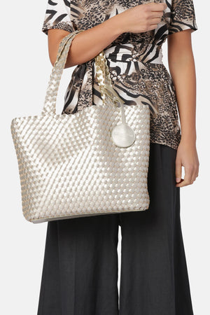 The WOVEN REVERSIBLE TOTE BAG by LINES OF DENMARK is a white tote featuring two shoulder straps and a circular faux-leather tag on one strap. This shopper showcases a textured, interlaced pattern and reversible design, providing stylish and elegant dual styling options. The image background is plain white.