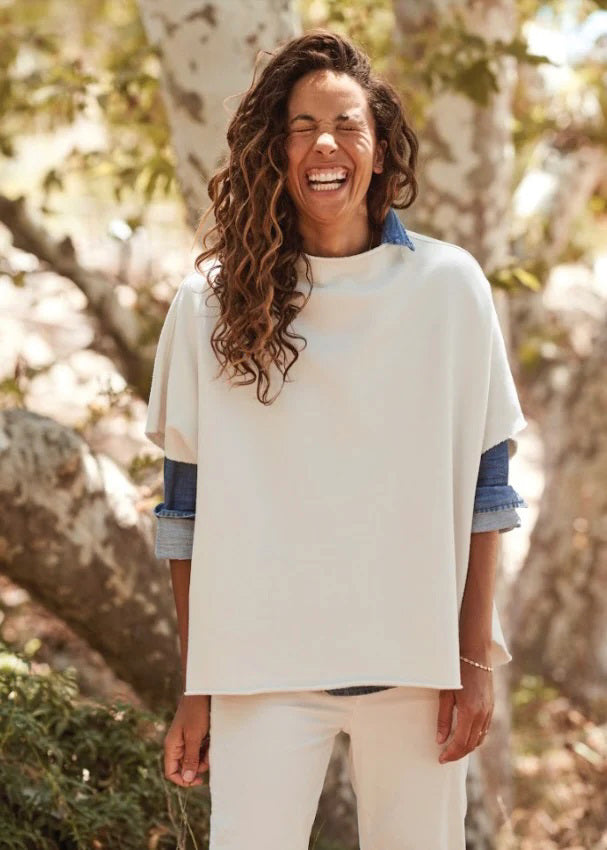 A person with long curly hair is laughing joyfully while wearing the Audrey Funnel Neck Capelet in Vintage White from Frank & Eileen over a blue shirt. They are standing outdoors in front of a tree, surrounded by sunlight and greenery.