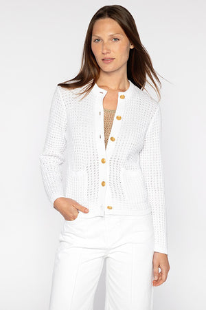 With long hair, a person wears the KINROSS CASHMERE TEXTURED BUTTONED CARDIGAN in white with gold accents over a beige top, paired with super soft cotton white pants. They stand against a plain white background with one hand in their pocket.