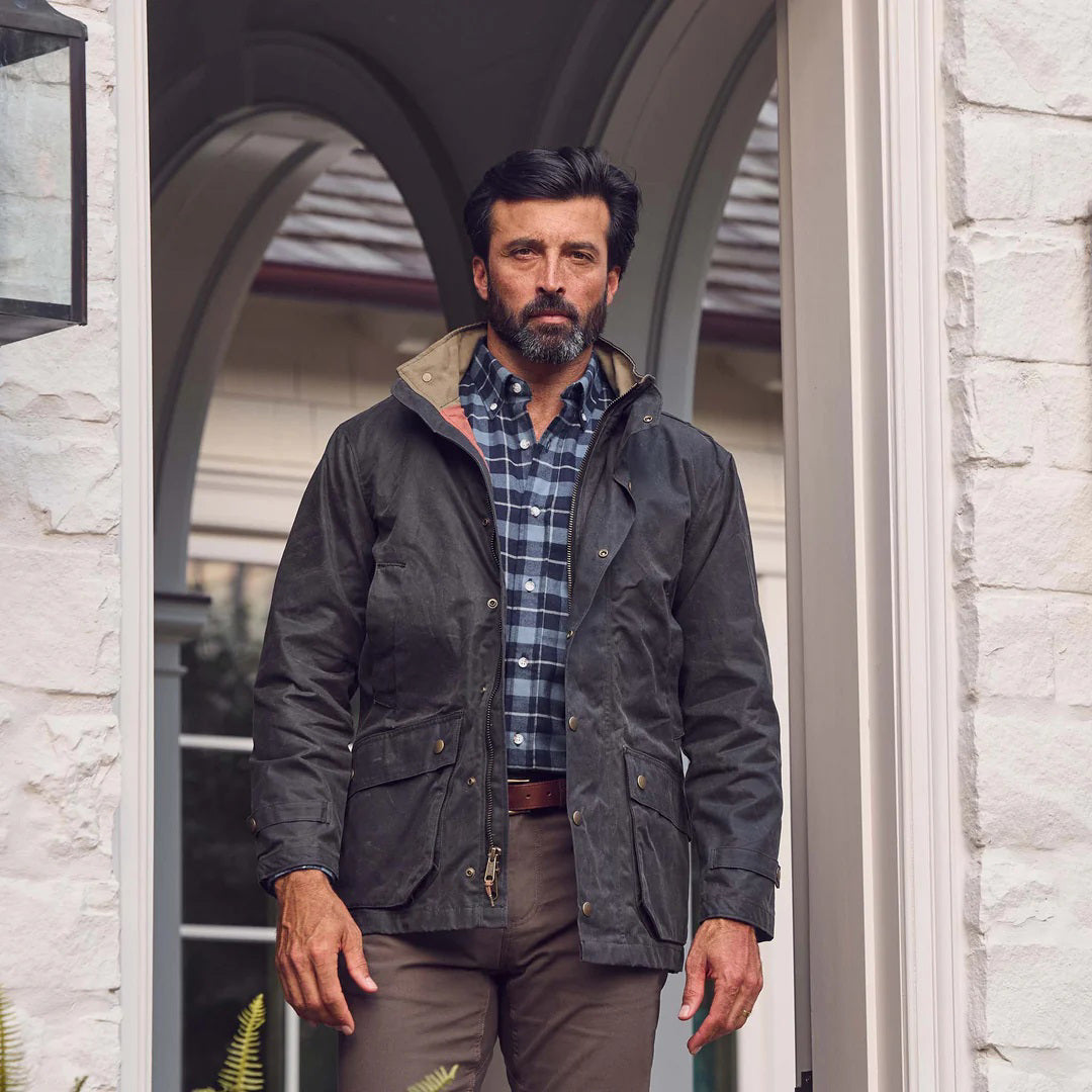 The TOM BECKBE Tenshaw Jacket in Hardwood is a dark green, USA-made waxed canvas jacket with a high collar, snap buttons, two front flap pockets, buttoned cuffs, and durable water-resistant shelter cloth for a rugged look.