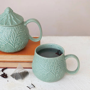 A textured green ceramic teapot and the EMBOSSED BOTANICALS MUG by CREATIVE COOP, both with ornate embossed patterns, sit on a stack of books. The mug is filled with tea, with a teabag nearby. The setting is on a light-colored surface, showcasing the sage reactive glaze beautifully.