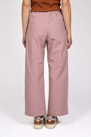 A person stands against a plain background wearing THE LITTLE PROJECT's AMBER PANT in pink, crafted from Japanese cotton poplin with a drawstring waist and subtle contrast stitching, paired with a sleeveless brown top and white and brown sneakers.