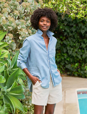 The FRANK & EILEEN Eileen Relaxed Button Up in Famous Denim Classic Wash is a light blue, long-sleeve shirt with bust-flattering buttons, a chest pocket, rolled sleeves, and a curved hem, shown against a white background.