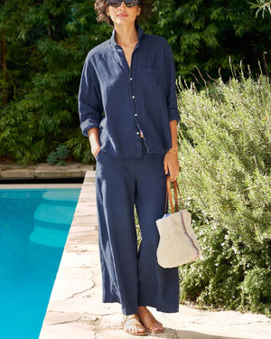 A person wearing a relaxed, navy blue button-up shirt and matching wide-leg silhouette pants from the FRANK & EILEEN Tuscany Getaway Set, including the FRANK AND EILEEN - MAISIE WIDE LEG LINEN PULL ON PANT, with hands in pockets, stands poolside on a sunny day. They carry a beige woven tote bag with blue and red stripes, have short curly hair, and wear sunglasses and sandals.