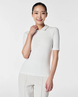 A woman in the SPANX - STRETCH RIB POLO TOP, a brown, ribbed, short-sleeve shirt with a button-down front, stands with her arms relaxed. The plain white background accentuates the top's stretchy ribbed fabric and body-hugging fit.