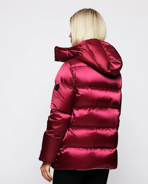 A person stands against a plain background, wearing the MIRTO 1956 - WATER REPELLENT QUILTED JACKET in a shiny red color, which features a removable hood. They are also dressed in black wide-leg pants and a light pink beanie. Their left hand hangs relaxed by their side while their right hand holds the jacket. Facing forward with a neutral expression, they complete the look effortlessly.