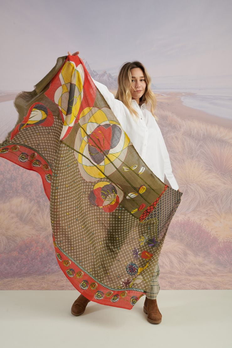 A woman with long blonde hair smiles at the camera in a scenic setting, wearing a white shirt, plaid pants, and the STORIATIPIC - ASTER WOOL SCARF by STORIATIPIC. Mountains, grass, and water are visible in the background.