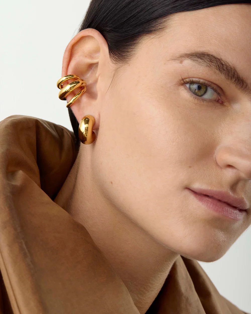 A close-up shows a person with dark hair pulled back and wearing a brown jacket, adorned with gold hoop earrings as well as wrap earrings featuring the JENNY BIRD - TOME TRIPLE EAR CUFF by JENNYBIRD. Crafted from 14k gold-dipped brass, these accessories shine brilliantly against the plain, light-colored background.