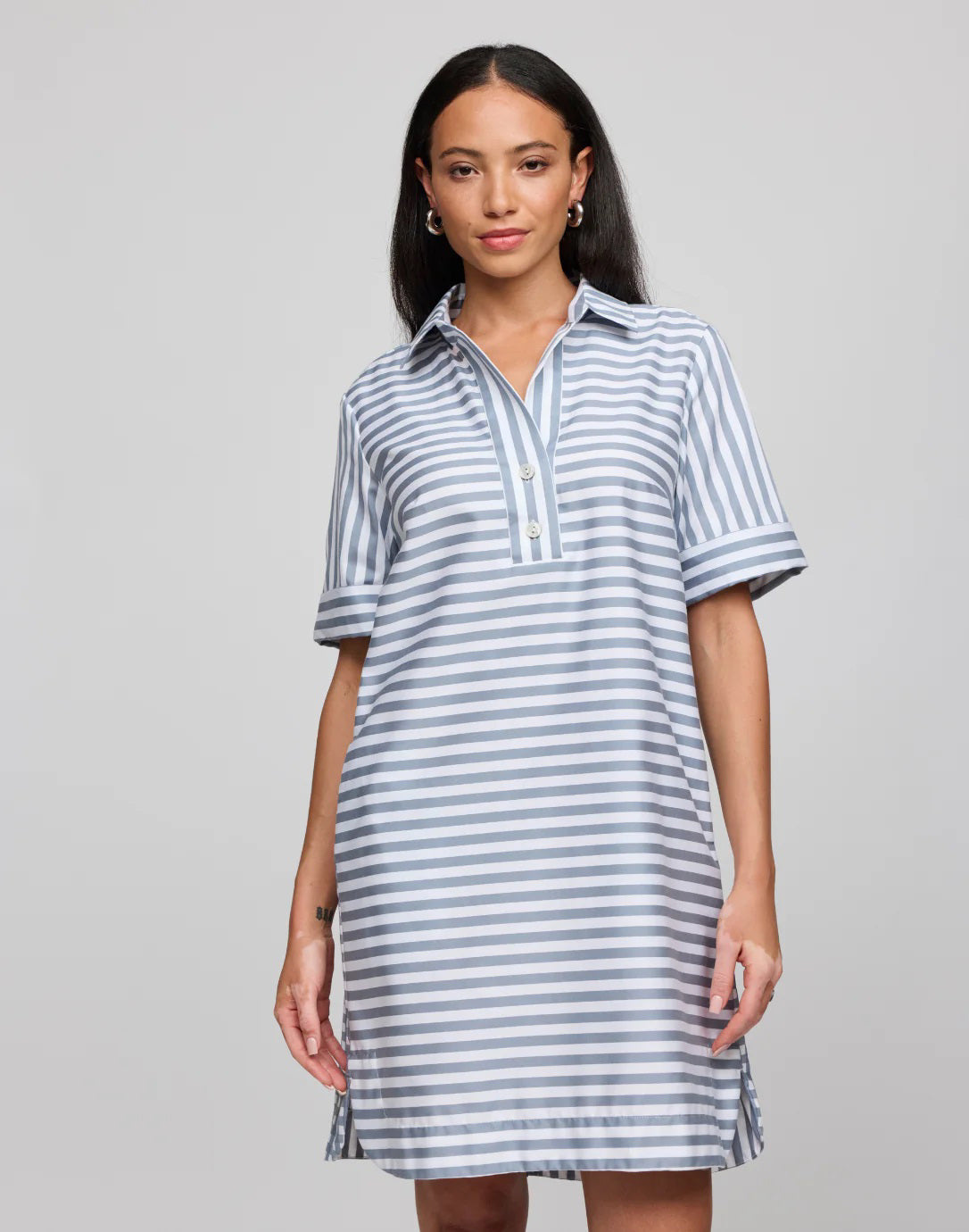 A woman in the HINSON WU Aileen Stripe and Gingham Dress, made from 100% cotton, stands against a plain backdrop. The dress features short sleeves and a collar. Her long dark hair cascades over her shoulders as she gazes directly at the camera.