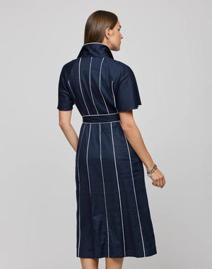 A woman with long brown hair wears the HINSON WU Joanna Dress, a navy knee-length piece featuring white piping and a waist tie belt made from stretch twill fabric, as she stands with hands in pockets against a plain background.