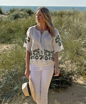 A woman with long blonde hair stands on a sandy beach with tall grass and the ocean in the background. She is wearing the ROSE & ROSE - ALASSIO TOP, a flattering empire line, white linen blend top adorned with daisy-inspired appliqué, paired with white pants and holding a straw hat.