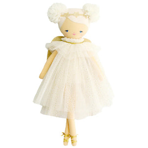 A young girl with light hair tied in a bow sits on a bed with white linens, smiling and holding up an Alimrose Ava Angel Doll Ivory Gold. She wears a light-colored dress, and behind her are pink and rust-colored pillows against a gray headboard.