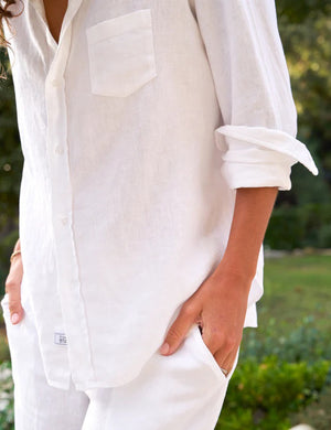 A woman with long hair is standing on a stone pathway outside, facing away from the camera. She is wearing the FRANK & EILEEN Eileen Relaxed Button-Up Shirt in Linen from the Tuscany Getaway Set paired with Maisie pull-on pants, both in a white, loose-fitting style. Her sleeves are rolled up and her hands are in her pockets. In the background, lush greenery and rocks reminiscent of a Tuscany Getaway set the scene.