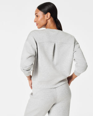 A woman stands confidently against a plain white background, wearing the SPANX Aireessentials Crew Neck Pullover and matching sweatpants made from luxurious comfort spacer fabric. She has her hair pulled back and her left hand is raised slightly. She smiles gently with her lips closed.