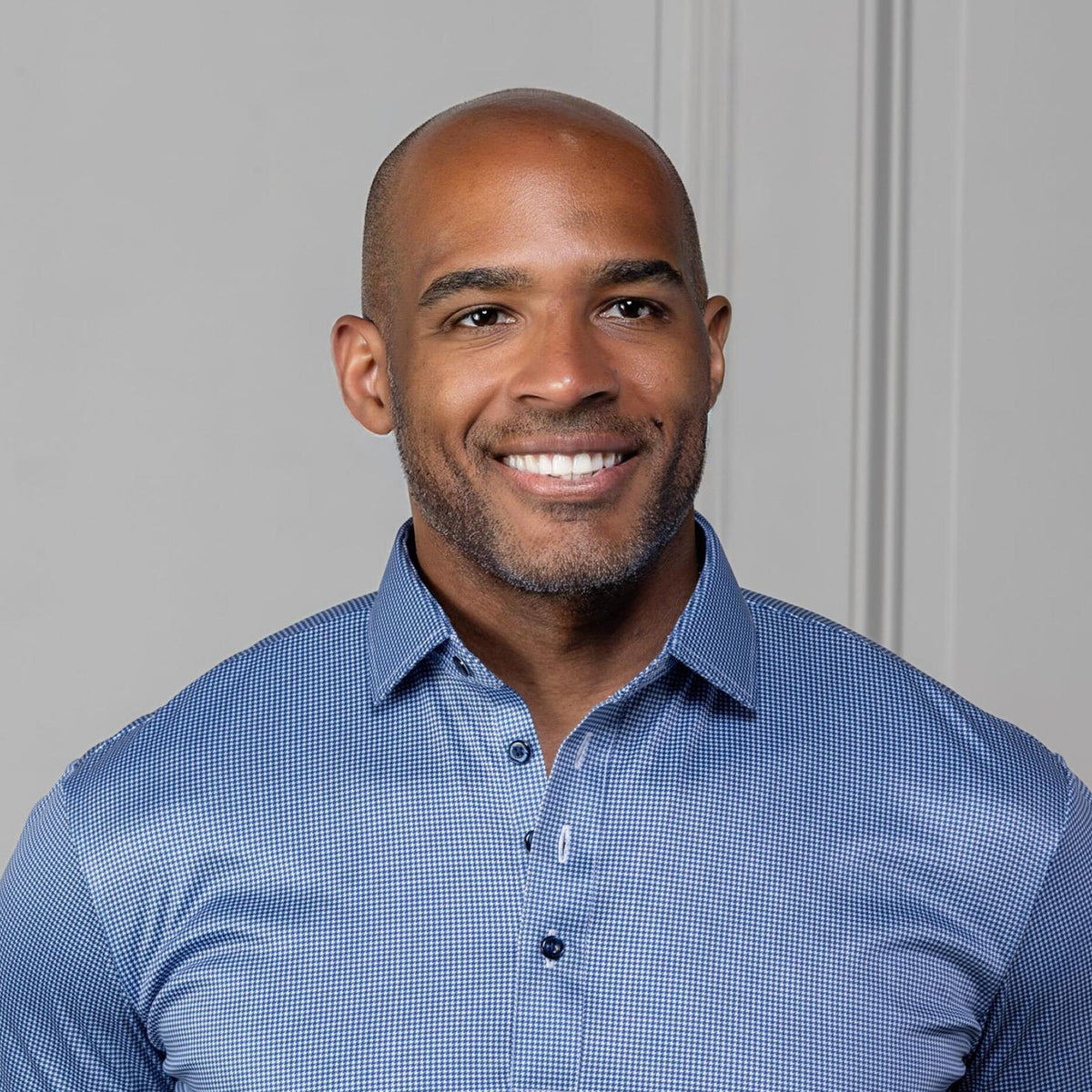 A man in sunglasses and a COLLARS AND CO - Semi Spread Collar Polo in Navy Houndstooth stands outdoors by a stone building and green plants, gazing to the side with one hand in his pocket.