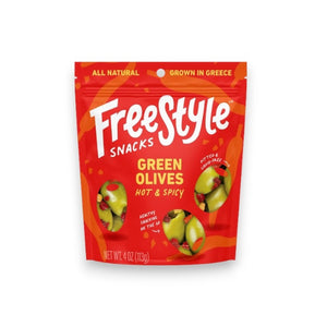 A vibrant orange snack bag labeled "FREESTYLE SNACKS" contains "FREESTYLE - GREEN OLIVES- HOT & SPICY," featuring spicy Calabrian chili. The 4 oz (113g) bag showcases images of green olives with red seasoning and highlights that the product is all-natural and grown in Greece.