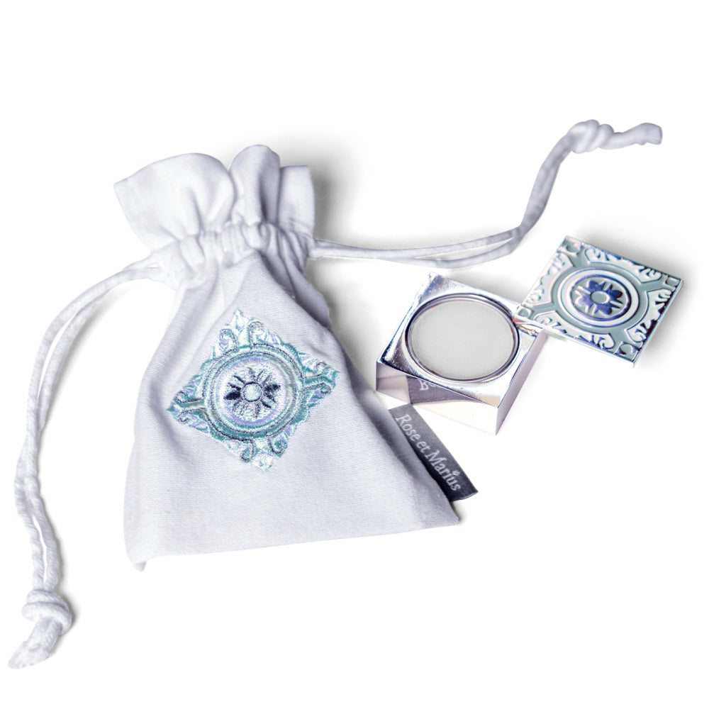 A manicured hand holds an open ROSE ET MARIUS - SUNLIT ROSE WATER SOLID PERFUME compact with a round pan, evoking subtle vanilla notes, against a white background.