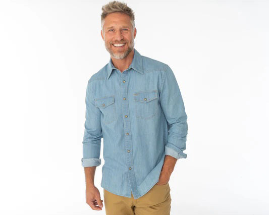 A man with a gray beard wears the CRIQUET WESTERN DENIM SHIRT WITH A PEARL SNAP IN LIGHT WASH by CRIQUET as he stands outdoors near a metal fence, with green grass and an open field in the background.
