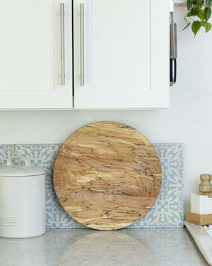 A small white vase filled with yellow and white daisy flowers sits on a PETERMANS BOARDS & BOWLS INC SPENCER PETERMAN - SPALTED LAZY SUSAN 20" along with a wedge of soft cheese topped with herbs and a few round crackers. The spinning tray rests on a kitchen counter, showcasing a patterned tile backsplash in the background.