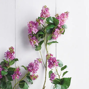 A sprig of vibrant purple GARDEN LILAC BRANCH 37 from NAPA HOME AND GARDEN, adorned with lush green leaves, rests against a plain white background. This showcases the stunning beauty of the blossoms and the striking contrast between their rich colors and the stark backdrop. Available for realistic portrayal, consider instore pickup to fully appreciate their charm up close.